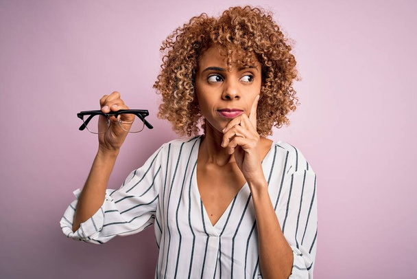 Afričan americká optická žena s kudrnaté vlasy drží brýle přes izolované růžové pozadí vážné tváře přemýšlení o otázce, velmi zmatený nápad - Fotografie, Obrázek