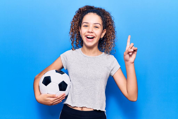 Красивая девочка с вьющимися волосами, держа футбольный мяч, удивлена идеей или вопросом, указывая пальцем со счастливым лицом, номер один  - Фото, изображение