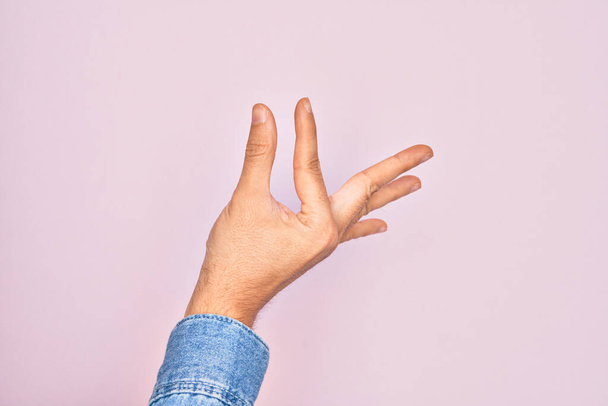 Dłoń białego młodzieńca pokazuje palce nad odizolowanym różowym tłem zbierając i biorąc niewidzialną rzecz, trzymając obiekt z palcami pokazującymi przestrzeń - Zdjęcie, obraz