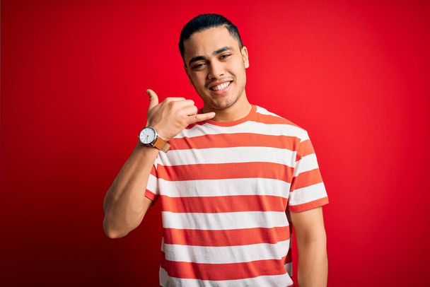 Joven brasileño con camiseta casual a rayas de pie sobre fondo rojo aislado sonriendo haciendo gesto telefónico con la mano y los dedos como hablando por teléfono. Comunicar conceptos
. - Foto, Imagen