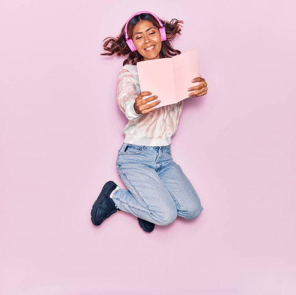 Молодая красивая латинская женщина слушает музыку в наушниках, улыбаясь счастливой. Прыжки с улыбкой на лице чтение книги на изолированном розовом фоне - Фото, изображение