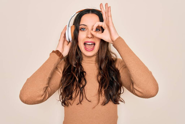 Young beautiful woman with blue eyes listening to music and dancing using headphones with happy face smiling doing ok sign with hand on eye looking through fingers - Foto, Bild
