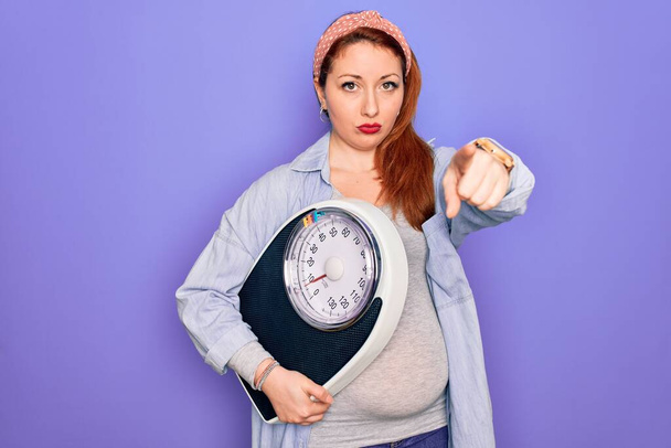 Young beautiful redhead pregnant woman expecting baby holding weighting machine pointing with finger to the camera and to you, hand sign, positive and confident gesture from the front - Photo, Image