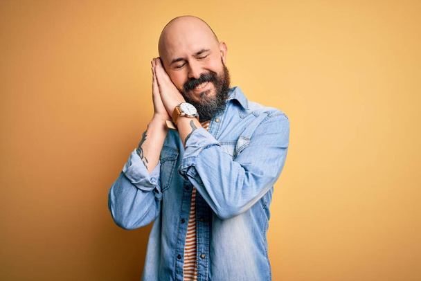 Handsome bald man with beard wearing casual denim jacket and striped t-shirt sleeping tired dreaming and posing with hands together while smiling with closed eyes. - 写真・画像