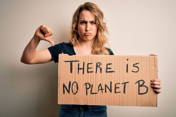 Jeune militante blonde demandant un environnement tenant une bannière avec un message planétaire avec un visage en colère, signe négatif montrant un aversion avec les pouces baissés, concept de rejet - Photo, image