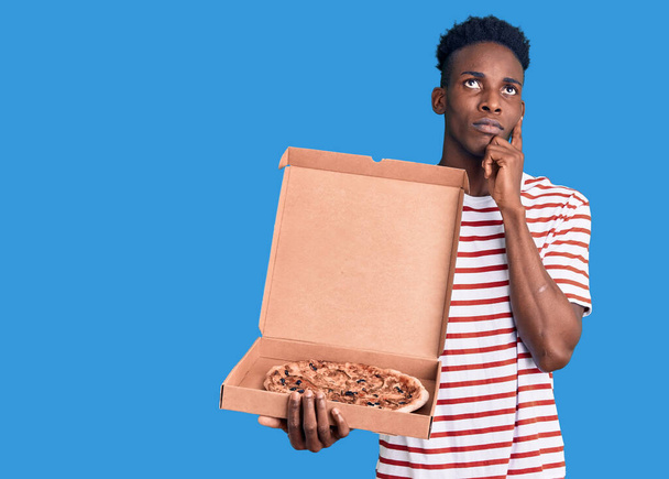 Jovem afro-americano segurando entrega caixa de pizza rosto sério pensando em questão com a mão no queixo, pensativo sobre ideia confusa  - Foto, Imagem