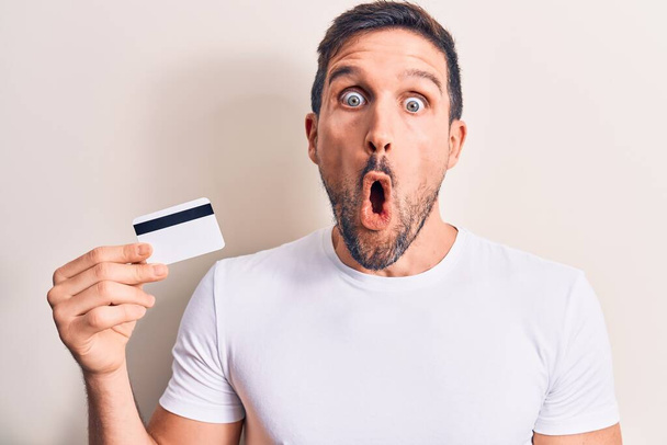 Junge gutaussehende Kunde Mann hält Kreditkarte über isolierten weißen Hintergrund verängstigt und erstaunt mit offenem Mund für Überraschung, ungläubiges Gesicht - Foto, Bild
