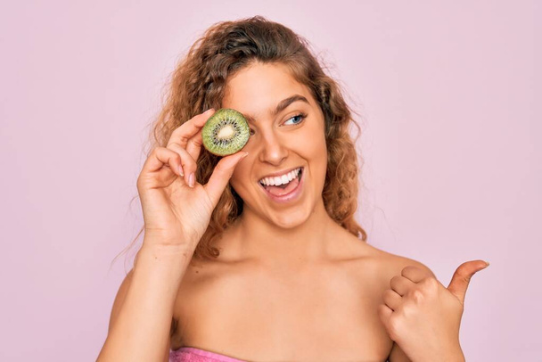 Beautiful woman with blue eyes wearing towel shower after bath holding kiwi fruit over eye pointing and showing with thumb up to the side with happy face smiling - Photo, Image