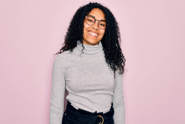 Młoda Afroamerykanka ubrana w sweter i okulary na różowym tle mruga do aparatu z seksownym wyrazem twarzy, radosną i szczęśliwą twarzą. - Zdjęcie, obraz