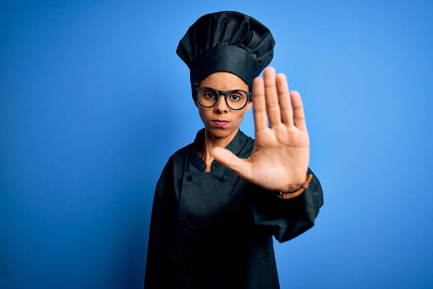 Giovane donna chef afroamericana indossa uniforme fornello e cappello su sfondo blu facendo smettere di cantare con il palmo della mano. Espressione di avvertimento con gesto negativo e serio sul viso. - Foto, immagini