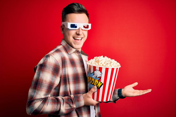 Joven hombre caucásico guapo usando gafas de película 3d y comer palomitas de maíz apuntando a un lado con las manos abiertas palmeras mostrando espacio de copia, presentando anuncio sonriendo emocionado feliz
 - Foto, Imagen