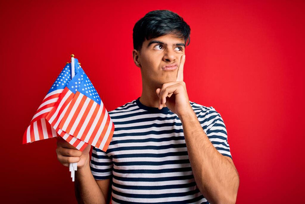 Молодой красивый патриот с флагами США празднует День независимости серьезное лицо, думая о вопросе, очень запутанная идея - Фото, изображение