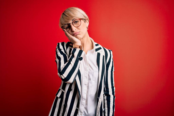 Blonde business woman with short hair wearing glasses and striped jacket over red background thinking looking tired and bored with depression problems with crossed arms. - Photo, image
