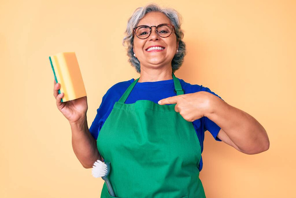 Senior hispanic grey- haired woman wearing apron holding scourer pointing finger to one self smiling happy and proud  - Photo, Image