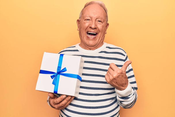 Senior handsome grey-haired man holding birthday gift over isolated yellow background pointing thumb up to the side smiling happy with open mouth - Foto, Imagem