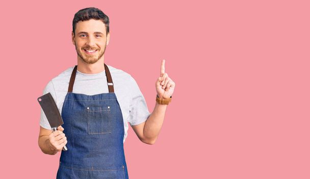 Handsome young man with bear wearing professional apron holding knife surprised with an idea or question pointing finger with happy face, number one  - Photo, Image