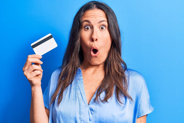 młoda piękna brunetka kobieta trzyma kartę kredytową na odizolowanym tle niebieski przestraszony i zdumiony z otwartymi ustami dla zaskoczenia, niedowierzanie twarz - Zdjęcie, obraz