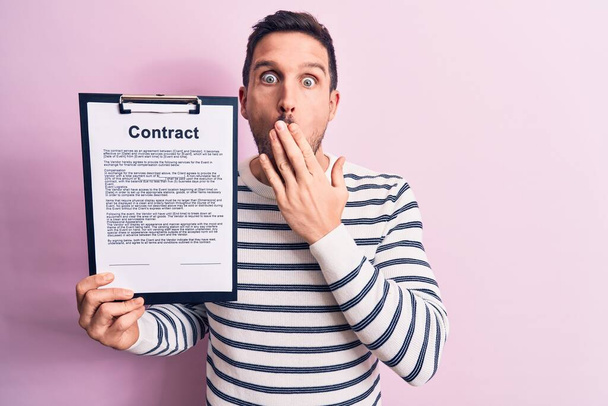 Young handsome man holding clipboard with contract document paper over pink background covering mouth with hand, shocked and afraid for mistake. Surprised expression - Zdjęcie, obraz