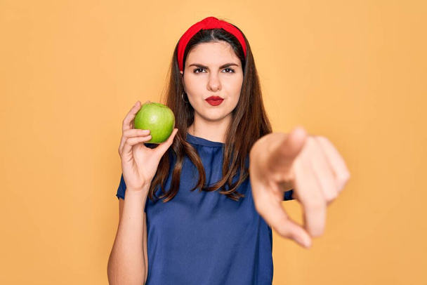 Молода красива дівчина їсть свіже органічне здорове зелене яблуко над жовтим тлом, вказуючи пальцем до камери і до вас, знак руки, позитивний і впевнений жест спереду
 - Фото, зображення