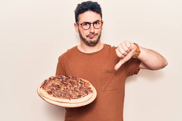 Молодой латиноамериканец в очках держит итальянскую пиццу с сердитым лицом, негативный знак показывает неприязнь с опущенными пальцами, концепция отказа  - Фото, изображение