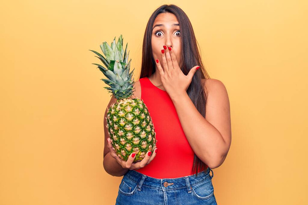 Nuori kaunis brunette nainen tilalla ananas peittää suun käsin, järkyttynyt ja pelkää virhe. yllättynyt ilme  - Valokuva, kuva