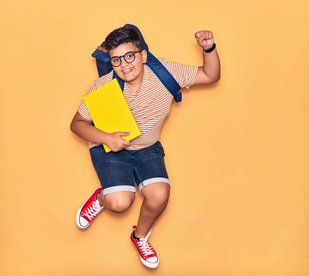 Adorable student boy wearing glasses and backpack smiling happy. Jumping with smile on face holding book celebrating with fist up over isolated yellow background. - Photo, image