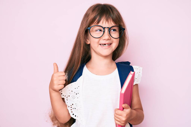 Kleines kaukasisches Mädchen mit langen Haaren, das einen Studentenrucksack trägt und ein Buch in der Hand hält, das glücklich und positiv lächelt, Daumen hoch und Zeichen der Anerkennung  - Foto, Bild
