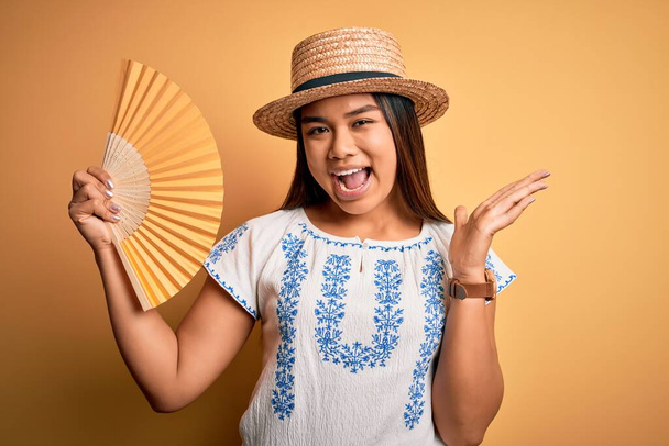 Junge asiatische Touristin im Urlaub trägt Sommermütze mit Handfächer über gelbem Hintergrund sehr glücklich und aufgeregt, Siegerausdruck feiert Sieg schreiend mit breitem Lächeln und erhobenen Händen - Foto, Bild