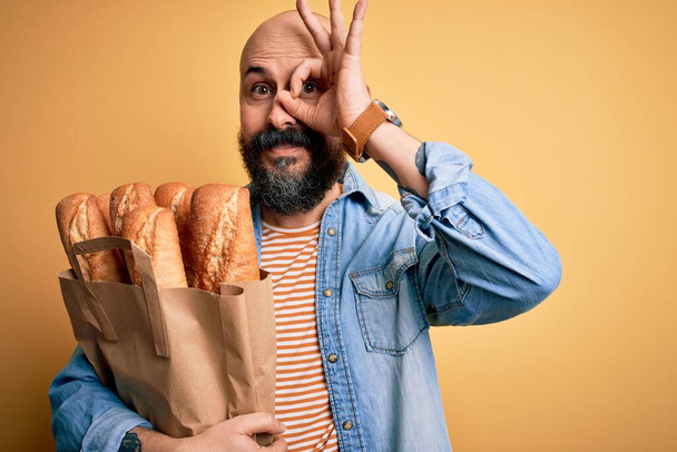 Jóképű kopasz férfi szakállal tartó papír táska kenyér sárga háttér boldog arc mosolygós csinál oké jel kéz a szemen néz keresztül ujjait - Fotó, kép