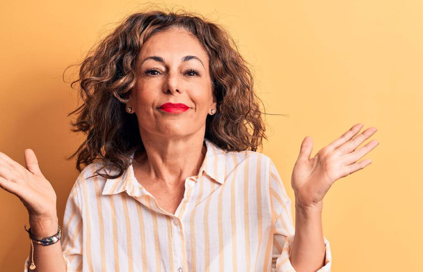 Middle age beautiful brunette woman wearing striped shirt standing over yellow background clueless and confused with open arms, no idea and doubtful face. - Photo, Image