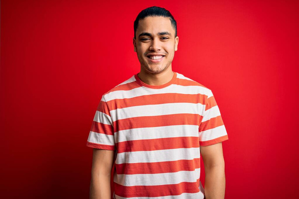 Молодой бразилец в повседневной полосатой футболке стоит на изолированном красном фоне со счастливой и прохладной улыбкой на лице. Счастливчик
. - Фото, изображение