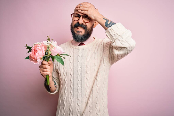 Handsome romantic bald man with beard holding beautiful bouquet over pink background stressed with hand on head, shocked with shame and surprise face, angry and frustrated. Fear and upset for mistake. - Photo, Image
