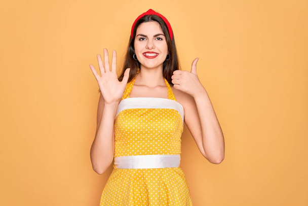 Nuori kaunis pin up nainen yllään 50s muoti vintage mekko keltainen tausta näyttää ja osoittaa sormilla numero kuusi hymyillen itsevarma ja onnellinen. - Valokuva, kuva