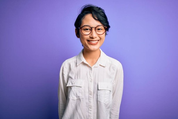 Young beautiful asian girl wearing casual shirt and glasses standing over purple background with a happy and cool smile on face. Lucky person. - Foto, Imagem