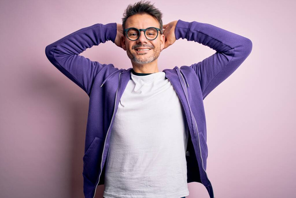 Молодий красивий чоловік у фіолетовому светрі та окулярах стоїть над рожевим тлом, розслабляючись та розтягуючись, руки та руки позаду голови та шиї усміхаються щасливо
 - Фото, зображення