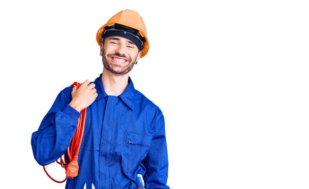 Jeune homme hispanique portant l'uniforme d'électricien tenant le câble regardant positif et heureux debout et souriant avec un sourire confiant montrant des dents  - Photo, image