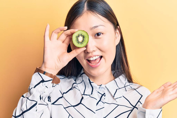 Mladá krásná čínská žena drží kiwi nad okem slaví úspěch s šťastným úsměvem a vítězný výraz se zvedl ruku  - Fotografie, Obrázek
