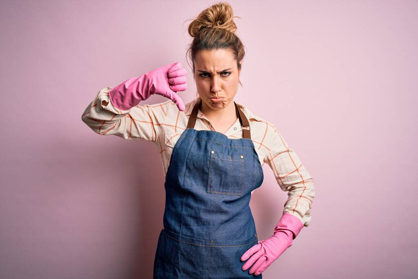 Young beautiful blonde cleaner woman doing housework wearing arpon and gloves looking unhappy and angry showing rejection and negative with thumbs down gesture. Bad expression. - Photo, Image