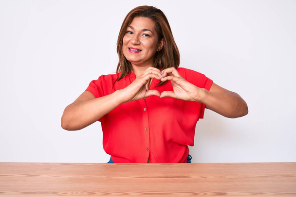 Keski-ikä brunette latino nainen yllään rento vaatteita istuu pöydällä hymyillen rakastunut tekee sydän symboli muoto kädet. romanttinen käsite.  - Valokuva, kuva