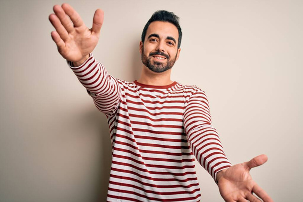 Young handsome man with beard wearing casual striped t-shirt standing over white background looking at the camera smiling with open arms for hug. Cheerful expression embracing happiness. - Photo, Image