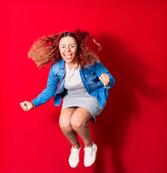Young beautiful latin woman wearing casual clothes smiling happy. Jumping with smile on face doing winner gesture with fists up over isolated red background - 写真・画像
