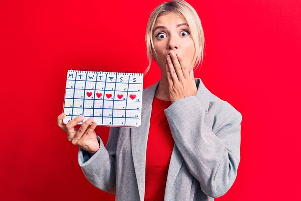 Bella donna bionda che controlla il calendario del periodo mestruale su sfondo rosso che copre la bocca con la mano, scioccata e spaventata per errore. Espressione sorpresa - Foto, immagini