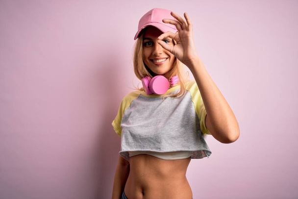 Młoda piękna blondynka sportowiec robi sport za pomocą słuchawek na różowym tle robi ok gest z uśmiechem dłoni, oko patrząc przez palce z szczęśliwą twarzą. - Zdjęcie, obraz