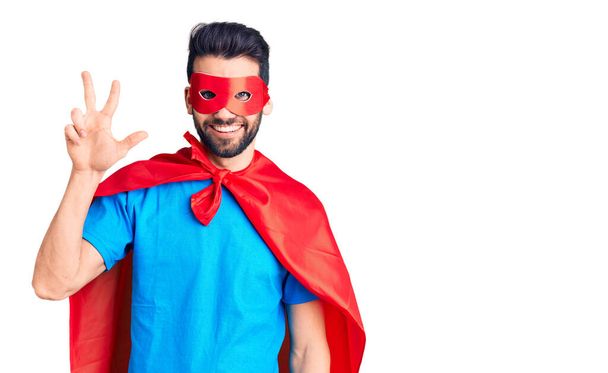 Young handsome man with beard wearing super hero costume showing and pointing up with fingers number three while smiling confident and happy.  - Photo, Image
