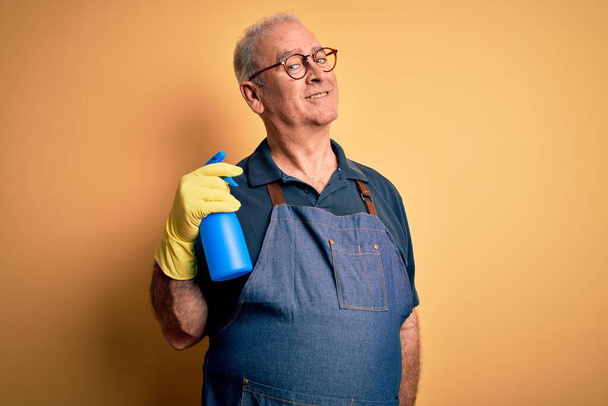 Middle age hoary cleaner man cleaning wearing apron and gloves using sprayer with a happy face standing and smiling with a confident smile showing teeth - Photo, Image