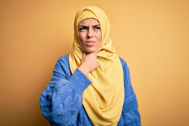Νεαρή όμορφη μελαχρινή μουσουλμάνα γυναίκα φορώντας αραβικό χιτζάμπ πάνω από απομονωμένο κίτρινο φόντο Αγγίζοντας επώδυνο λαιμό, πονόλαιμο για γρίπη, θρόμβο και λοίμωξη - Φωτογραφία, εικόνα