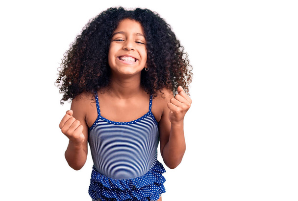 Afrykańskie amerykańskie dziecko z kręconymi włosami noszące stroje kąpielowe bardzo szczęśliwe i podekscytowane robiąc zwycięski gest z podniesionymi ramionami, uśmiechnięte i krzyczące o sukces. koncepcja uroczystości.  - Zdjęcie, obraz