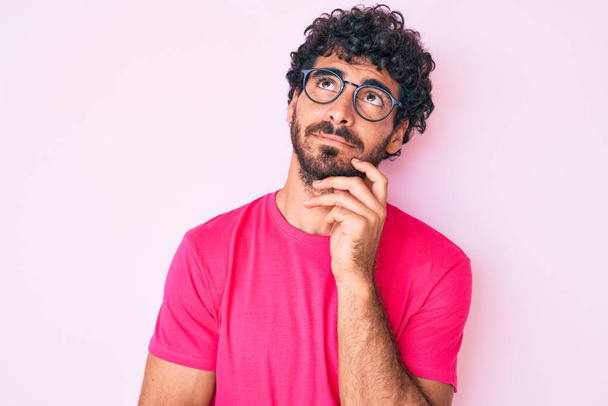 Handsome young man with curly hair and bear wearing casual clothes and glasses serious face thinking about question with hand on chin, thoughtful about confusing idea  - Photo, Image