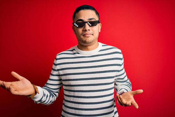 Young brazilian man wearing funny thug life sunglasses over isolated red background clueless and confused expression with arms and hands raised. Doubt concept. - 写真・画像