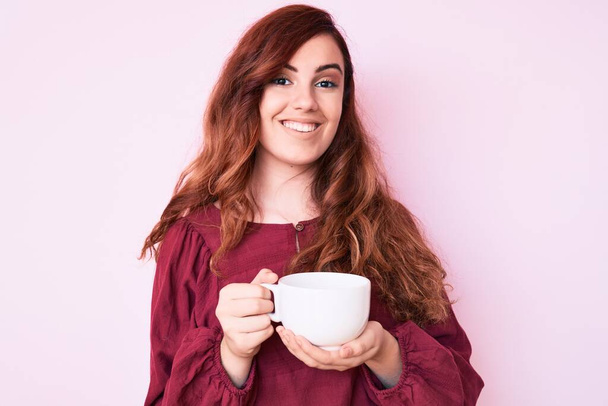 Молодая красивая женщина держит кофе глядя позитивно и счастливо стоя и улыбаясь с уверенной улыбкой показывая зубы  - Фото, изображение